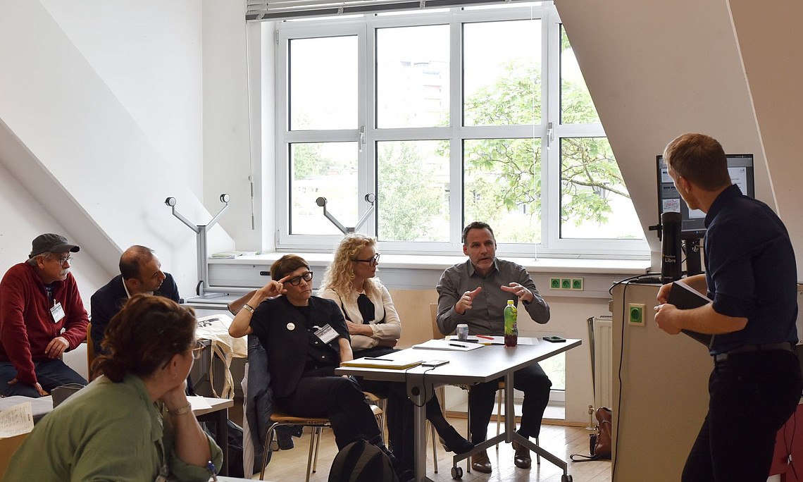 6 Leute sitzen an Tischen in einem Seminarraum und sprechen mit einem Mann, der am Rednerpult steht.