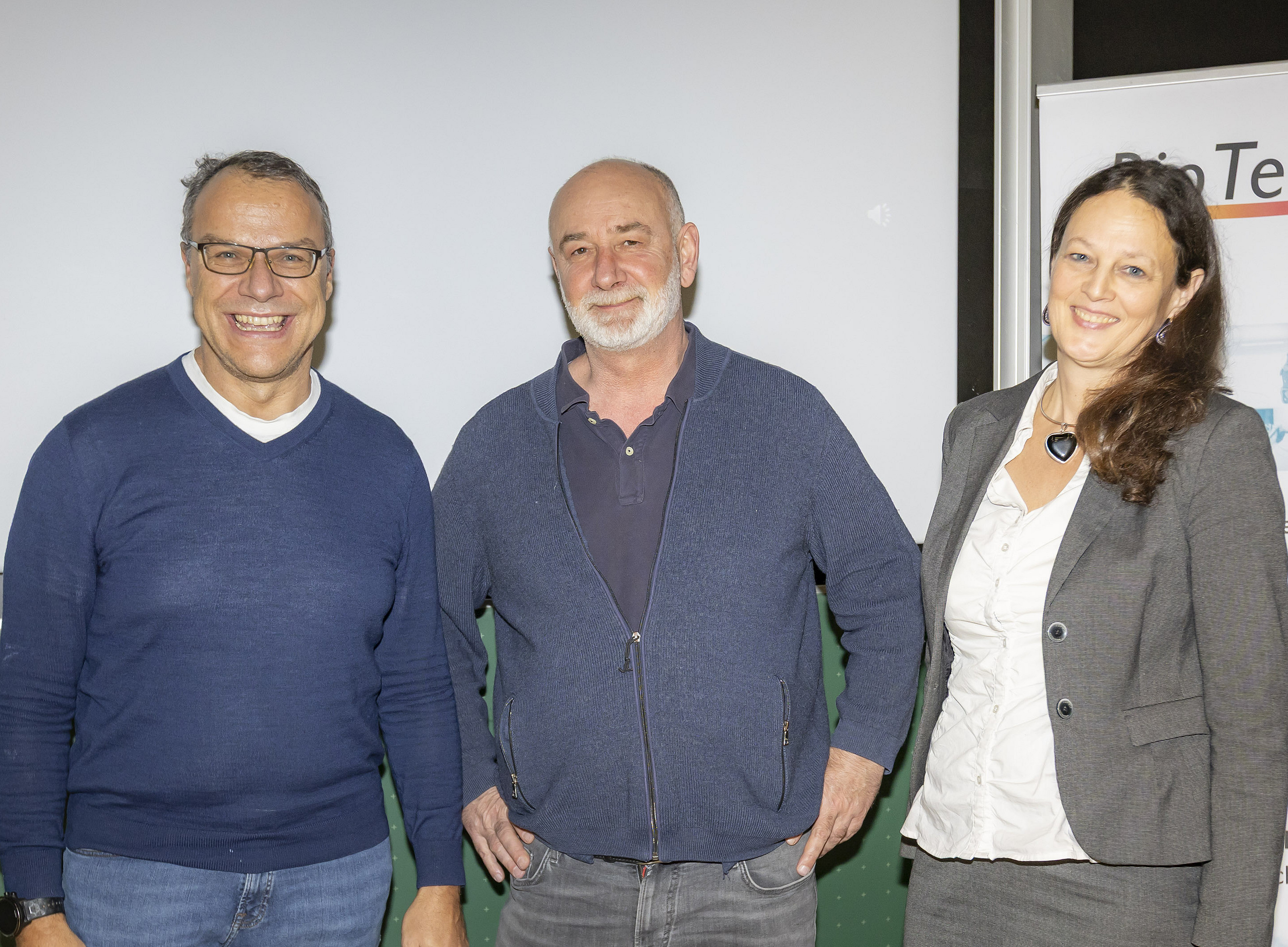 Three people are standing side by side, smiling at the camera. On the left is a man wearing glasses and a blue jumper; in the middle is a man with a grey beard and a blue cardigan; and on the right is a woman with long brown hair, wearing a grey suit and a white blouse. In the background, a white screen and a roll-up banner with partially visible lettering can be seen. ©Foto Fischer, Graz