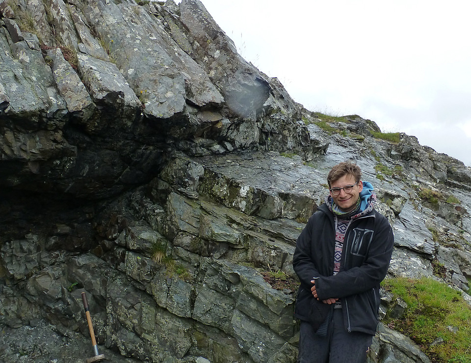 Fotografie von Geowissenschaften-Absolvent Arthur Borzi im Freien vor einem Felsen, an dem ein Hammer lehnt.