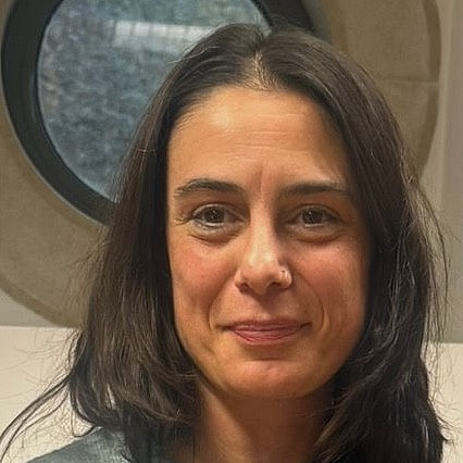 Das Foto zeigt Raquel Carvalheira, eine Frau mit langen Haaren, die ihr Kinn auf ihre Hand stützt, während sie sitzt und eine Jeansjacke trägt. Im Hintergrund sind verschwommene Figuren und sanftes Licht zu sehen, was auf eine Innenraumkulisse hindeutet. ©R. Baldé