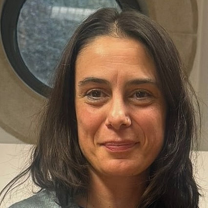 Das Foto zeigt Raquel Carvalheira, eine Frau mit langen dunklen Haaren, die einen hellgrauen Cardigan über einem blauen Hemd trägt und vor einem runden Fenster steht und direkt in die Kamera blickt. ©R. Baldé