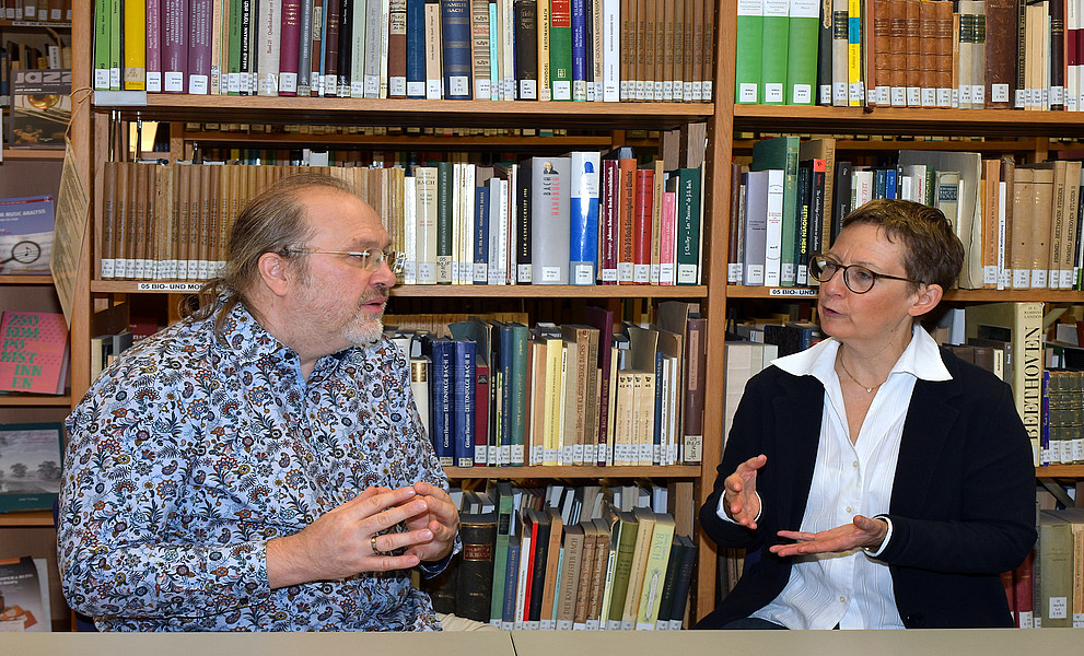 Stefan Baumgarten und Susanne Kogler im Gespräch, an einem Tisch sitzend vor einem Bücherregal