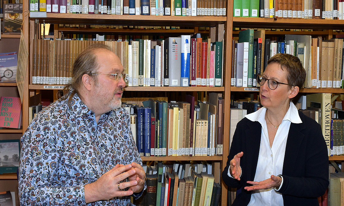 Stefan Baumgarten und Susanne Kogler im Gespräch, an einem Tisch sitzend vor einem Bücherregal