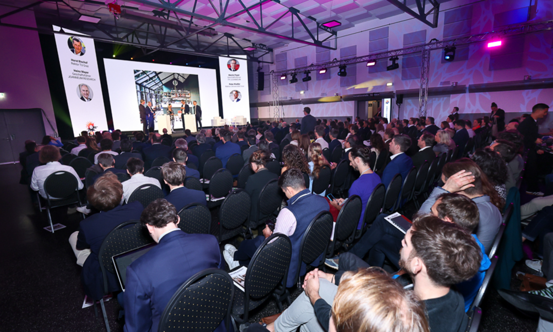 The main stage of the conference featured a young audience in suits, sitting and standing. Screens displayed images about business innovation in the field of AI. The large hall had modern architecture and was illuminated by purple lights. 