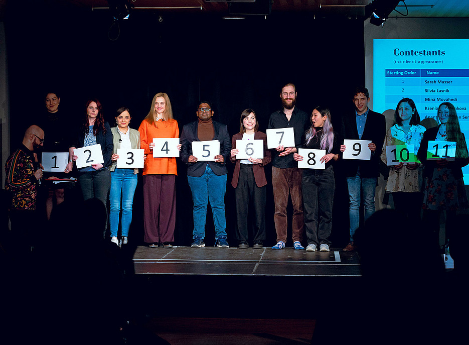Die elf 3MT-Teilnehmer:innen der Uni Graz nebeneinander, mit jeweils einer Tafel mit der Nummer ihrer Platzierung ©Uni Graz/Angele