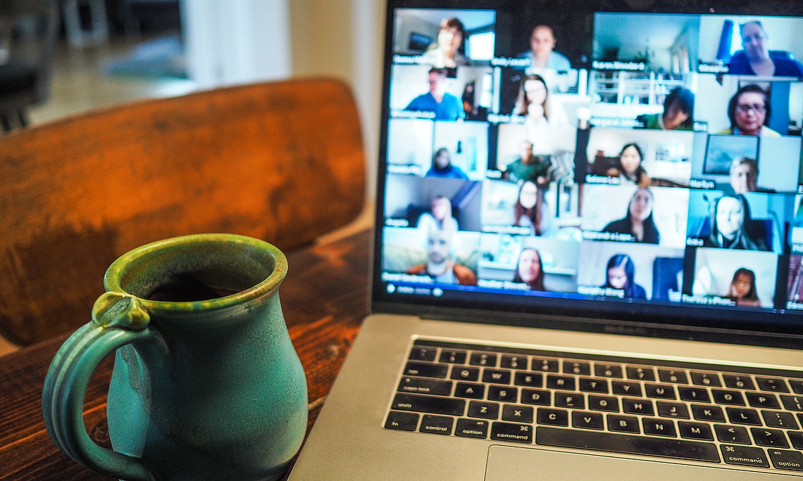 Laptop auf einem Schreibtisch mit einer türkisfarbenen Tasse daneben und auf dem Bildschirm ist eine Gruppe von Personen in Form einer Galerie in einem online Meeting zu sehen.