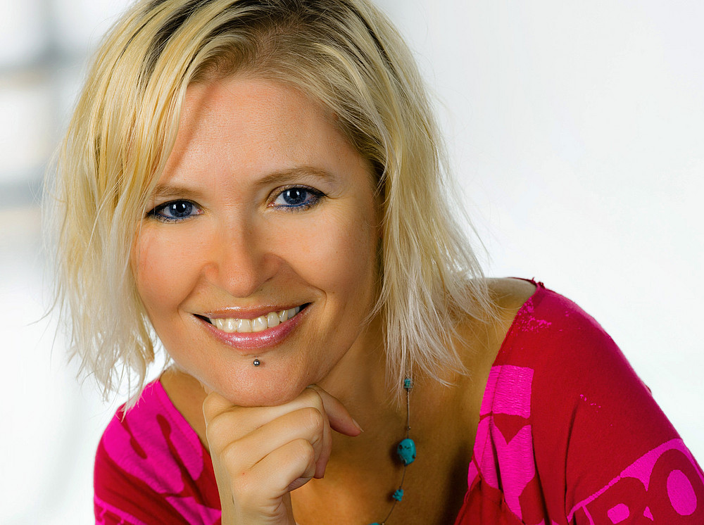 Porträt einer Frau mit schulterlangen blonden Haaren und einem Unterlippen-Piercing. Sie trägt ein roten Shirt und hat ihren Kopf auf eine Hand gestützt. ©Foto Wilke