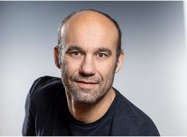 Mittelalter Mann mit hoher Stirn, kurzem Haar und Bartansatz trägt dunkles T‑Shirt, schaut frontal in die Kamera vor neutral grauem Hintergrund. ©© Helmut Lunghammer