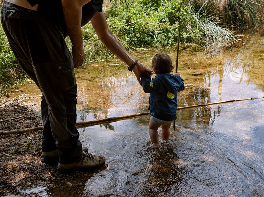 The photo shows an adult holding the hand of a child standing in shallow water, surrounded by greenery and reflections of trees in the water. The child is wearing a blue jacket. ©Sylvain Maestraggi