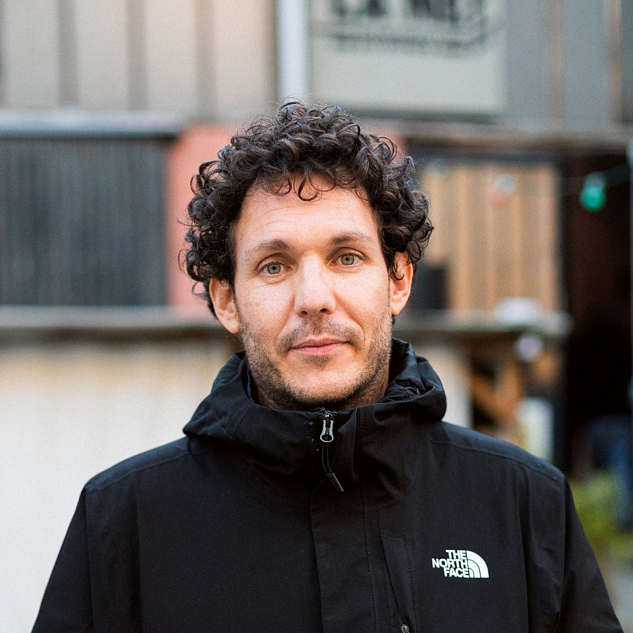 Das Foto zeigt Samuel Sami Everett, einen Mann mit lockigem Haar, der eine schwarze North Face-Jacke trägt und im Freien steht. Im Hintergrund ist ein rustikales Gebäude mit Metallverkleidung zu sehen. ©Elodie Martial