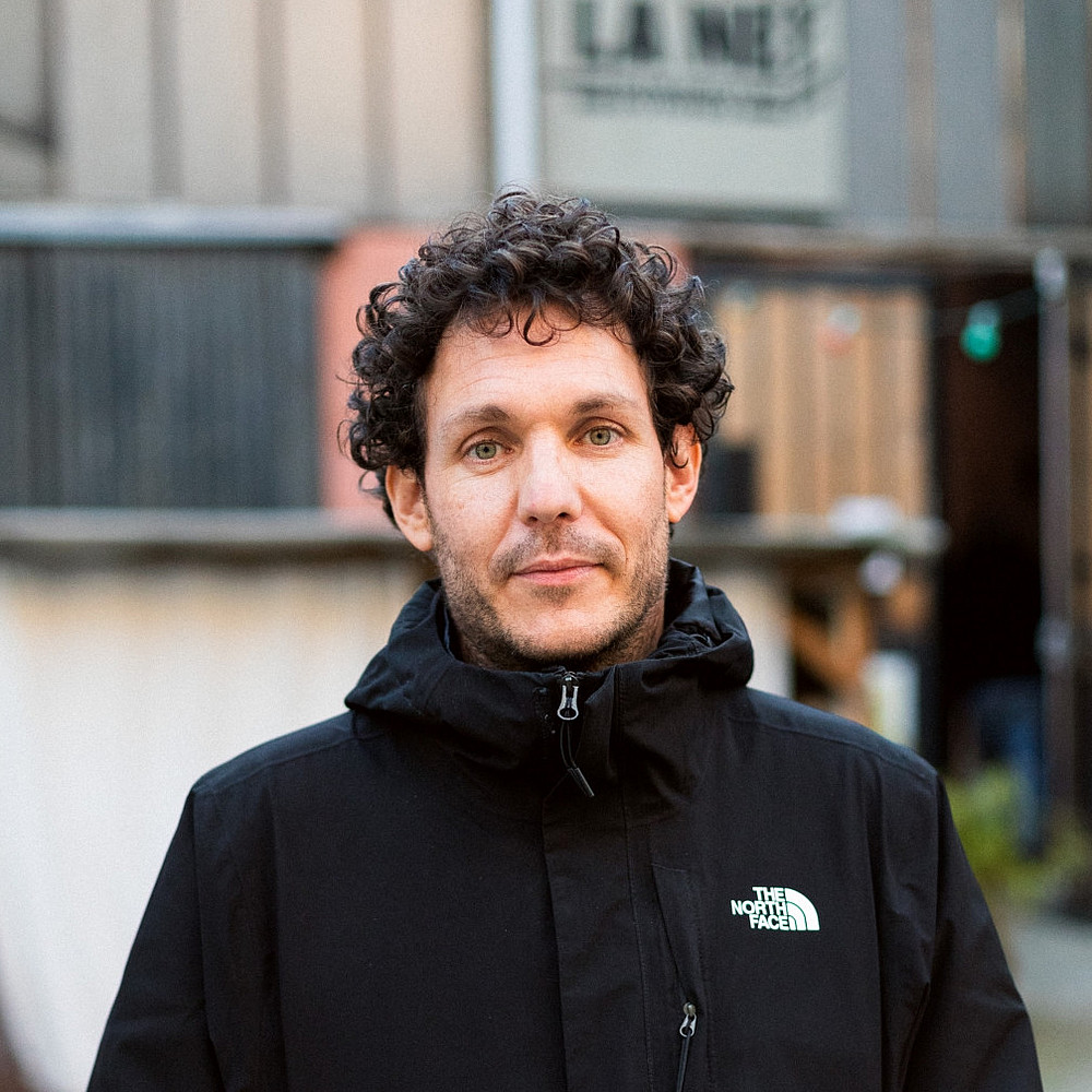 Ein Porträt von Samuel Sami Everett, einem Mann mit lockigem Haar, der eine schwarze North Face-Jacke trägt, steht im Freien. Im Hintergrund ist ein rustikales Gebäude mit Metallverkleidung zu sehen. ©Elodie Martial