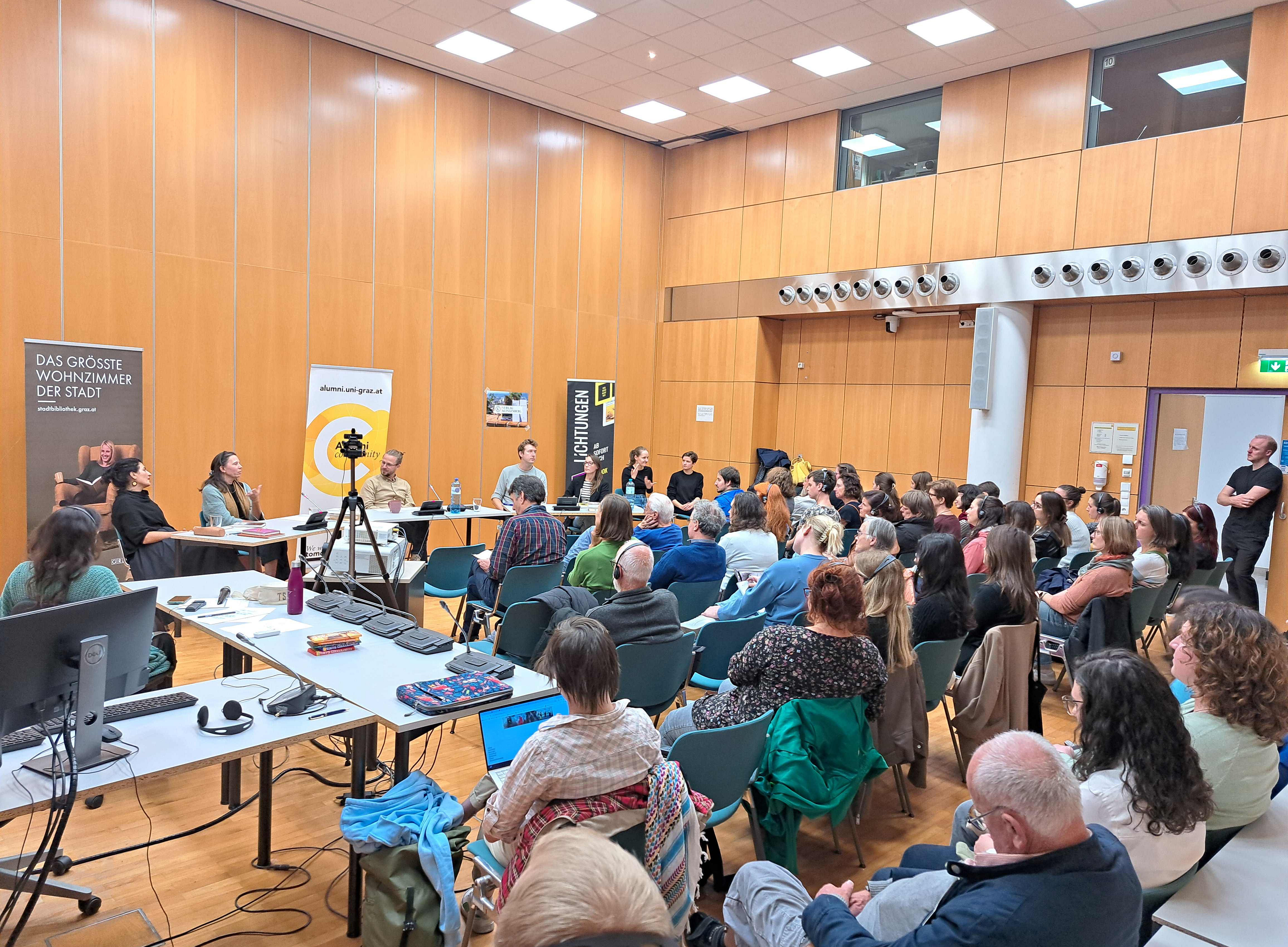 Personen sprechen vor vollbesetztem Saal; Blick von vorne 