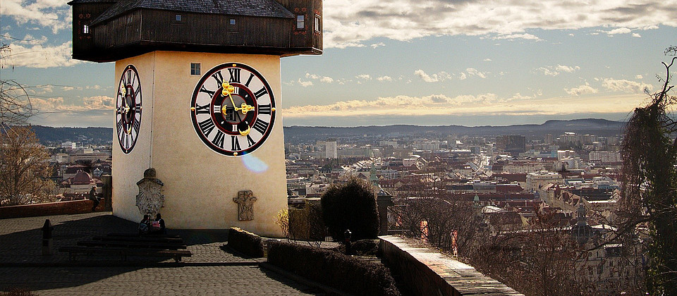 Blick auf den Grazer Uhrturm und dahinter die Dächer der Stadt bei Sonnenschein.