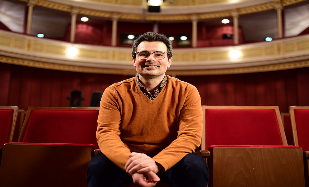 Ein Mann mit einem orangen Pullover und Brille sitzt in einem festlichen Saal mit roter Samt-Bestuhlung Saal. 