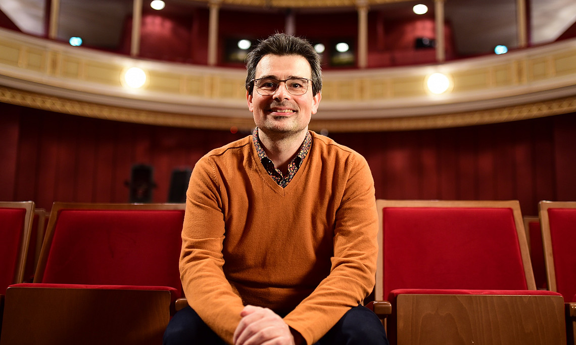 Ein Mann mit einem orangen Pullover und Brille sitzt in einem festlichen Saal mit roter Samt-Bestuhlung Saal. 