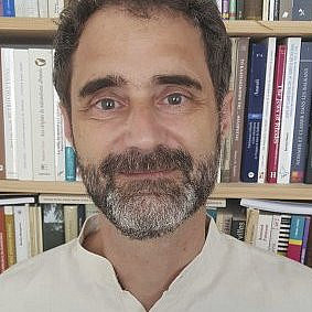 Das Foto zeigt Pierre Sintes, einen Mann mit Bart und kurzen Haaren, der ein helles Hemd trägt und vor einem Bücherregal voller verschiedener Bücher steht. ©Pierre Sintes