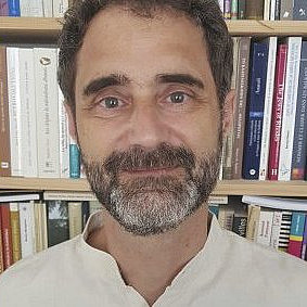 Das Bild zeigt Pierre Sintes, einen Mann mit Bart und Schnurrbart, der vor einem Bücherregal voller verschiedener Bücher lächelt. Er trägt ein helles Hemd mit Kragen. ©Pierre Sintes