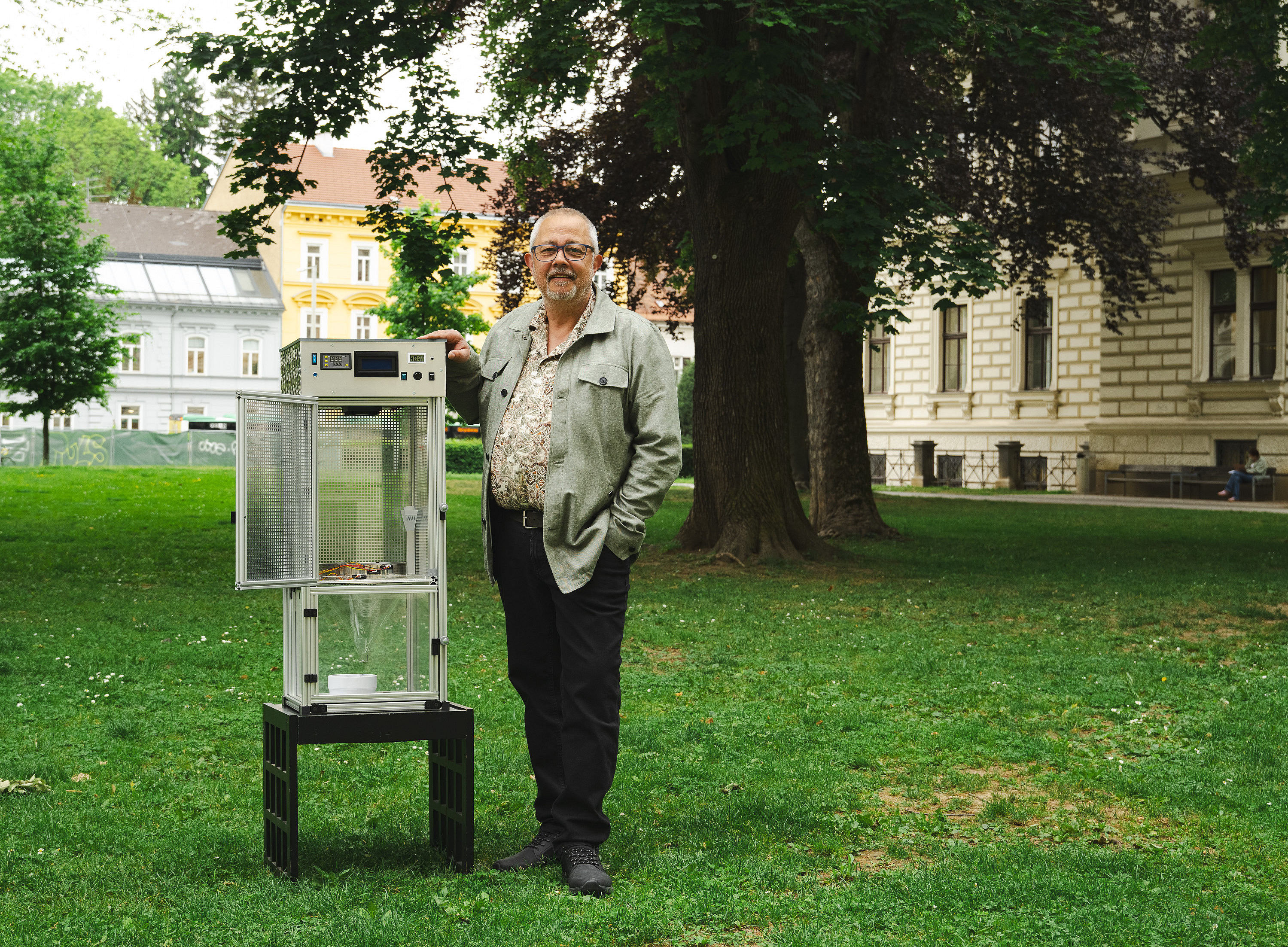 Ein Mann mittleren Alters steht neben einer offenen Glasvitrine auf der Wiese eines Parks auf dem Campus der Universität Graz. Die Vitrine hat er unter den Arm geklemmt. Er lächelt leicht in die Kamera und posiert für ein Foto ©Uni Graz/Tzivanopoulos