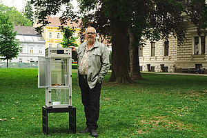 Ein Mann mittleren Alters steht neben einer offenen Glasvitrine auf der Wiese eines Parks auf dem Campus der Universität Graz. Die Vitrine hat er unter den Arm geklemmt. Er lächelt leicht in die Kamera und posiert für ein Foto