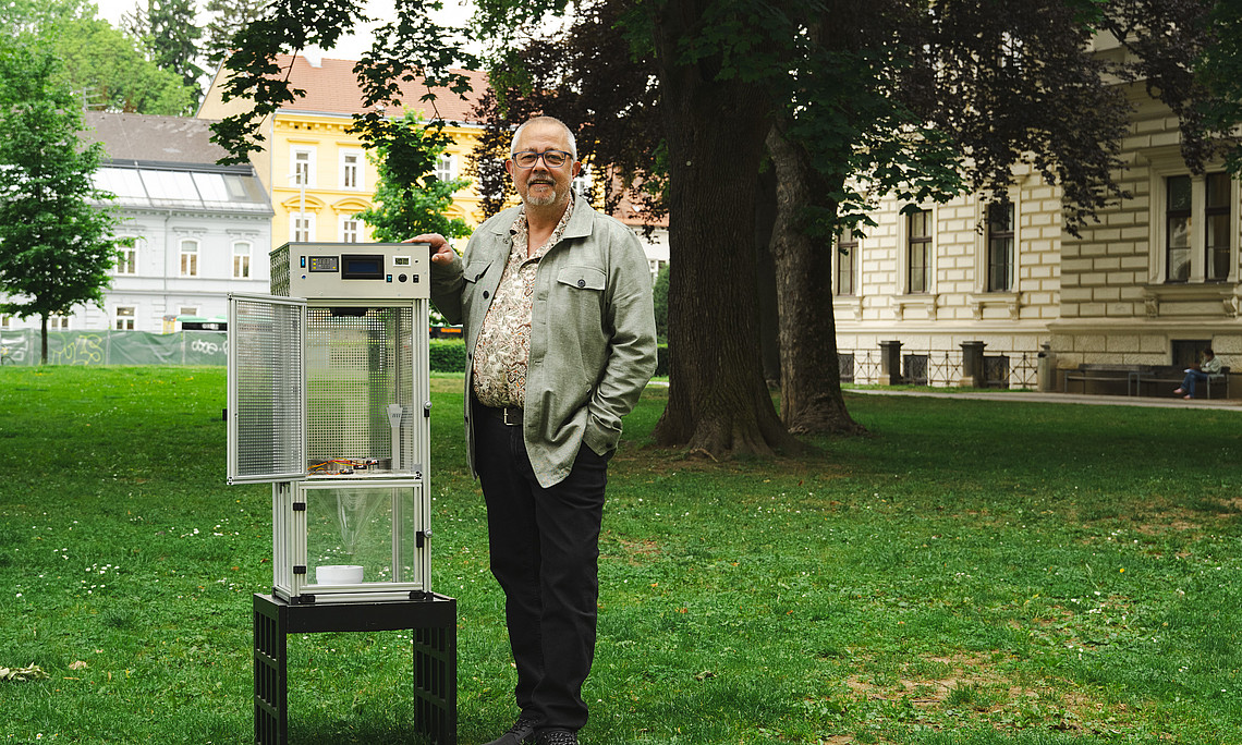 A middle-aged man stands next to an open glass cabinet on the lawn of a park on the Campus of the University of Graz. The cabinet is placed under his arm. He smiles slightly at the camera and poses for a photo