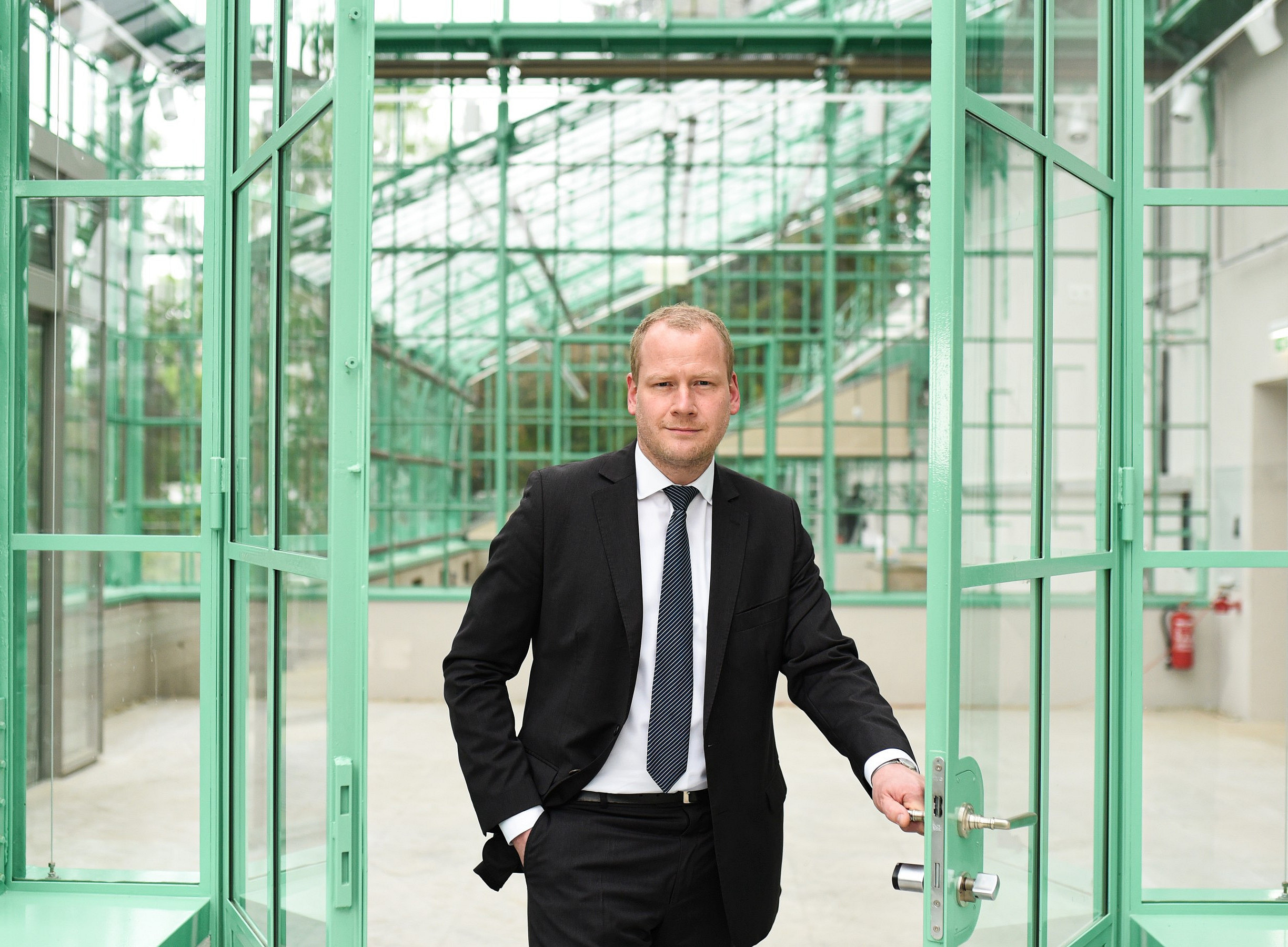 Porträt eines Geschäftsmannes im Anzug, der die Tür zu einem Gebäude mit grünem Glas, einem großen industriellen Eisenrahmen und einer Stahlkonstruktion offen hält. Er steht in einem Innengarten. 