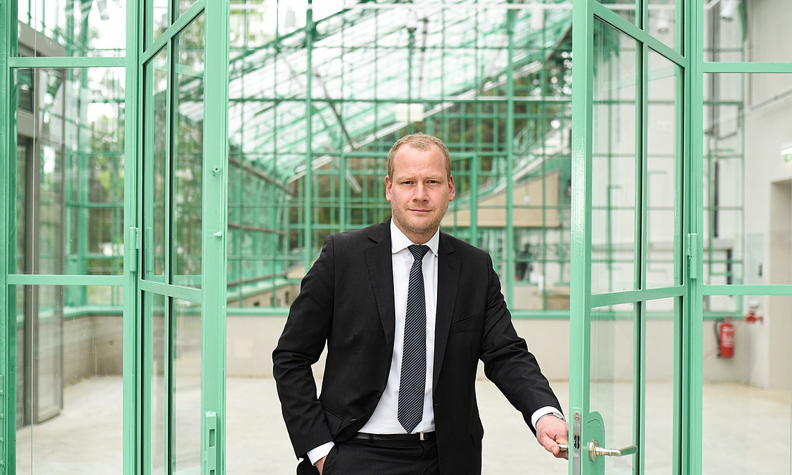 Portrait of a businessman in a suit, holding open the door to a green glass entrance building with a large industrial iron frame and steel structure. he is standing inside an indoor garden