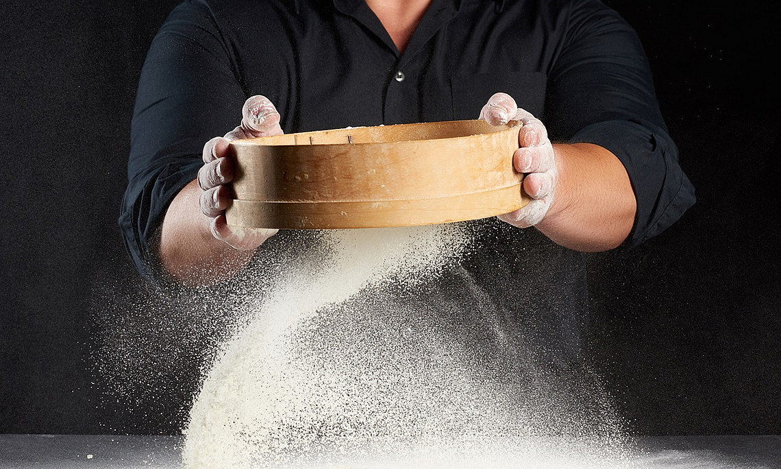 Mann vor schwarzem Hintergrund, der ein Sieb hält, aus dem weißes Mehl nach unten fällt