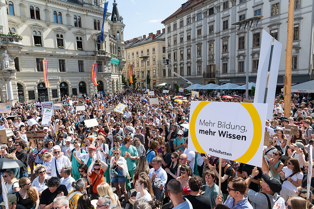 Große Menschenmenge unterschiedlichen Alters demonstriert in sonniger Innenstadt, viele halten Plakate mit Texten zu Forschung, Bildung und Gesellschaft hoch. ©Uni Graz/Angele