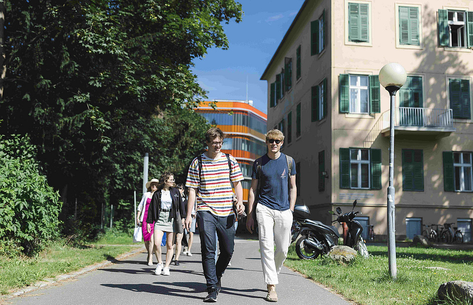 Zwei junge Männer flanieren einen Weg am Campus der Uni Graz entlang, Studierenden vor dem Hauptgebäude der Uni Graz, Sujetbild fürs Studium Uni Graz