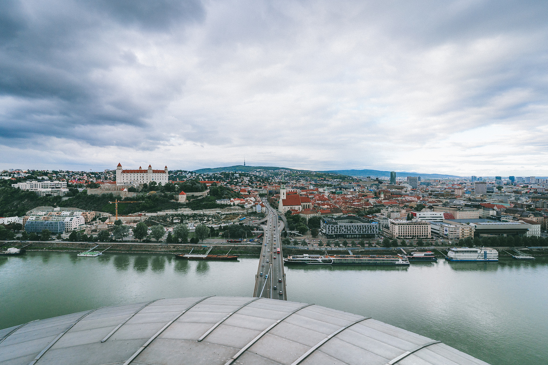 Panoramic view of a European city along a wide river under a cloudy sky. A bridge stretches across the water toward a historic old town with red-roofed buildings and a prominent castle perched on a hill to the left. Church spires rise among dense city blocks, while modern buildings stand farther in the background. Boats are docked along the riverbanks, and the foreground shows the curved roof of a structure overlooking the scene. ©Manuel Madaini