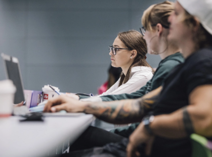 Foto von Universitätsstudenten, die an Schreibtischen in einem Hörsaal sitzen und an Laptops arbeiten. Eine Studentin hat Tattoos und trägt eine Brille. ©Universität Graz/Kanizaj