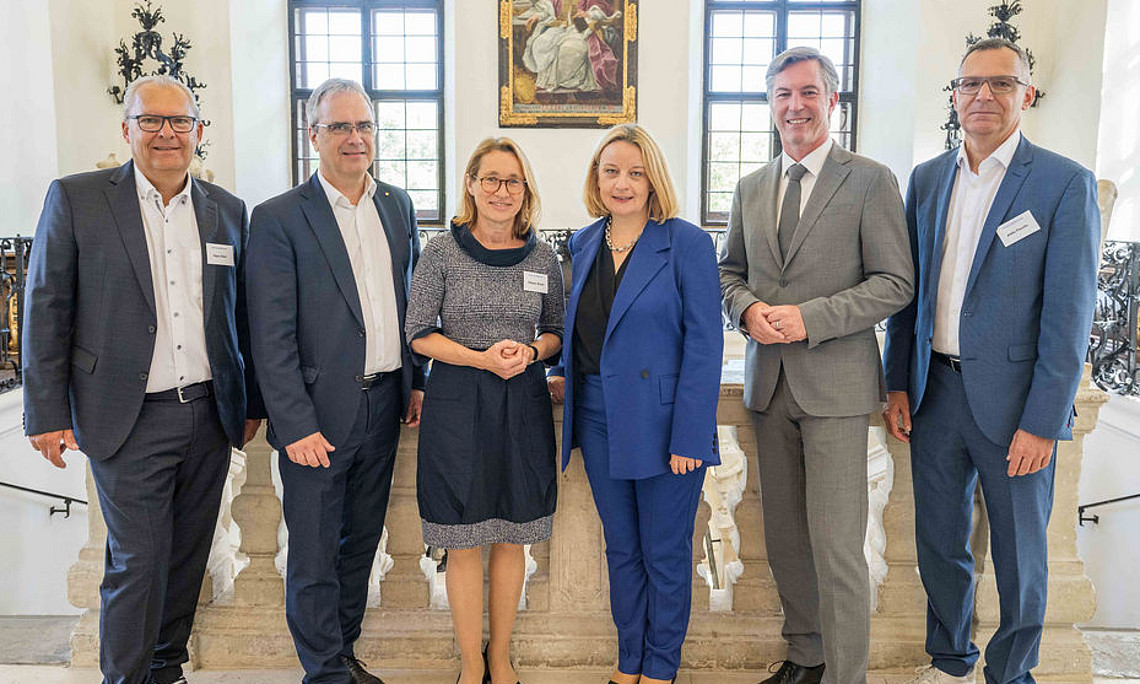 a group photo of a university summit with three men and two women standing next to each other smiling at camera, wearing business attire. 