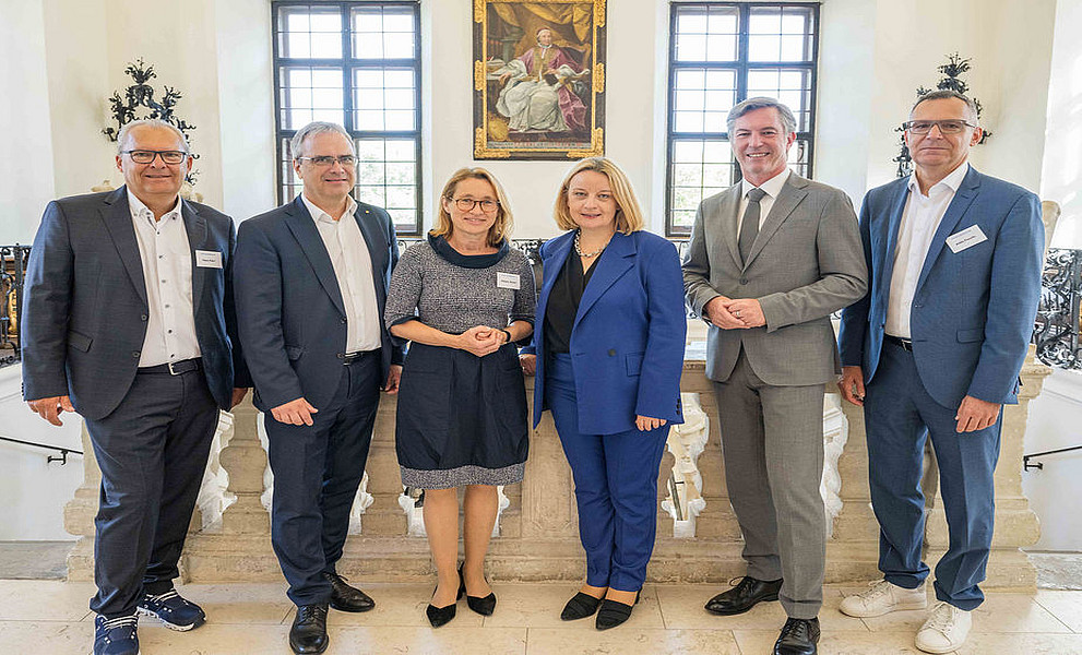 Ein Gruppenfoto von einem Universitätsgipfel mit drei Männern und zwei Frauen, die nebeneinander stehen, in die Kamera lächeln und Business-Kleidung tragen. 
