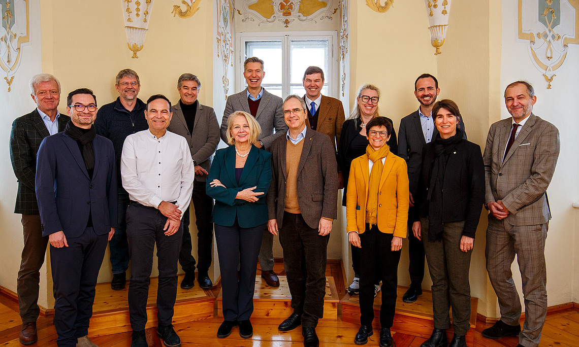 Gruppenfoto von 14 Personen in Business- und Smart-Casual-Kleidung in einem historischen Innenraum mit verzierten Stuckelementen, hellen Wänden und Holzparkett. Die Gruppe steht auf einem erhöhten Holzpodest vor einem Fenster; vorne mehrere Personen mit verschränkten Armen oder Händen vor dem Körper, rechts eine Person in grauem Anzug, links Personen in Jacken und Schals. Die Atmosphäre wirkt formell und freundlich