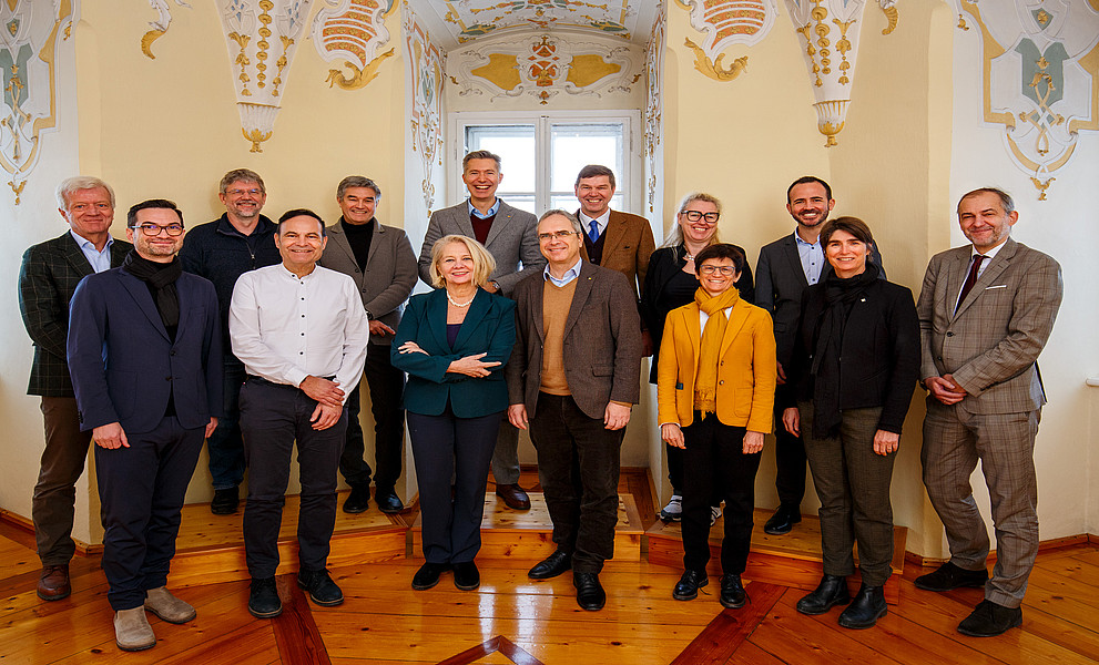 Gruppenfoto von 14 Personen in Business- und Smart-Casual-Kleidung in einem historischen Innenraum mit verzierten Stuckelementen, hellen Wänden und Holzparkett. Die Gruppe steht auf einem erhöhten Holzpodest vor einem Fenster; vorne mehrere Personen mit verschränkten Armen oder Händen vor dem Körper, rechts eine Person in grauem Anzug, links Personen in Jacken und Schals. Die Atmosphäre wirkt formell und freundlich