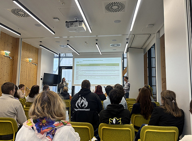 zwei Frauen stehen vor einem Tisch mit Laptop und halten einen Vortrag, hinter ihnen sieht man die Leinwand mit der Präsentation. Im Publikum sitzen viele Menschen und hören zu ©Uni Graz/Schmieder