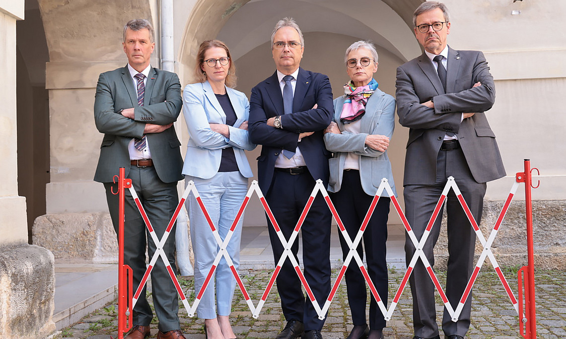 Vier Rektor:innen und eine Vizerektorin stehen mit verschränkten Armen und finsteren Gesichtern hinter einer Absperrung.