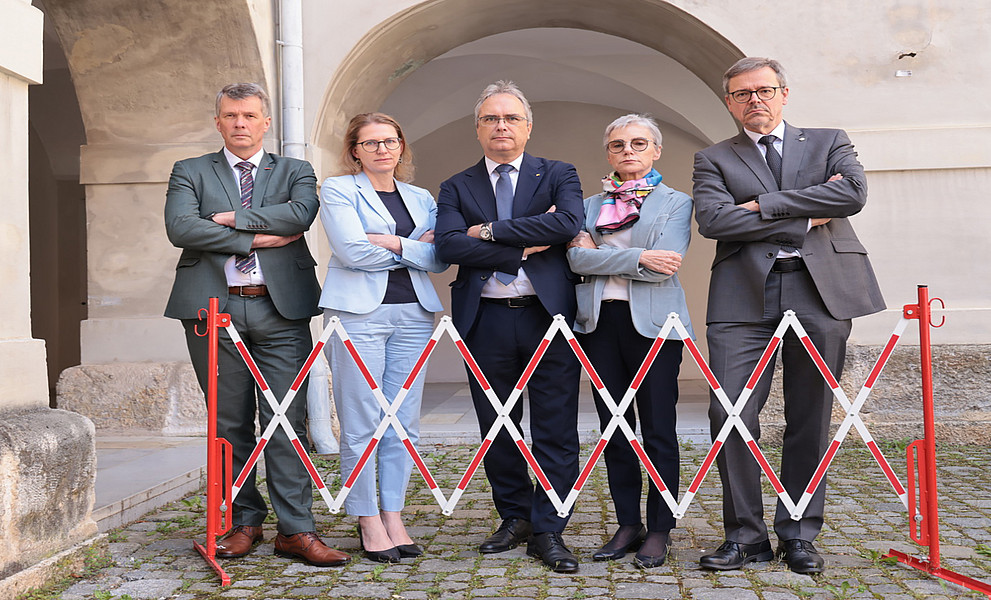 Vier Rektor:innen und eine Vizerektorin stehen mit verschränkten Armen und finsteren Gesichtern hinter einer Absperrung.