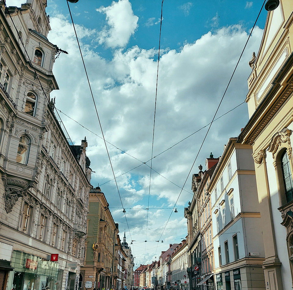 Einkaufsstraße in der Grazer Altstadt, in der Mitte sind Schienen für die Straßenbahn verlegt, rechts und links säumen kleine Geschäft in alten Gebäuden die Straße; vereinzelt sind Menschen unterwegs