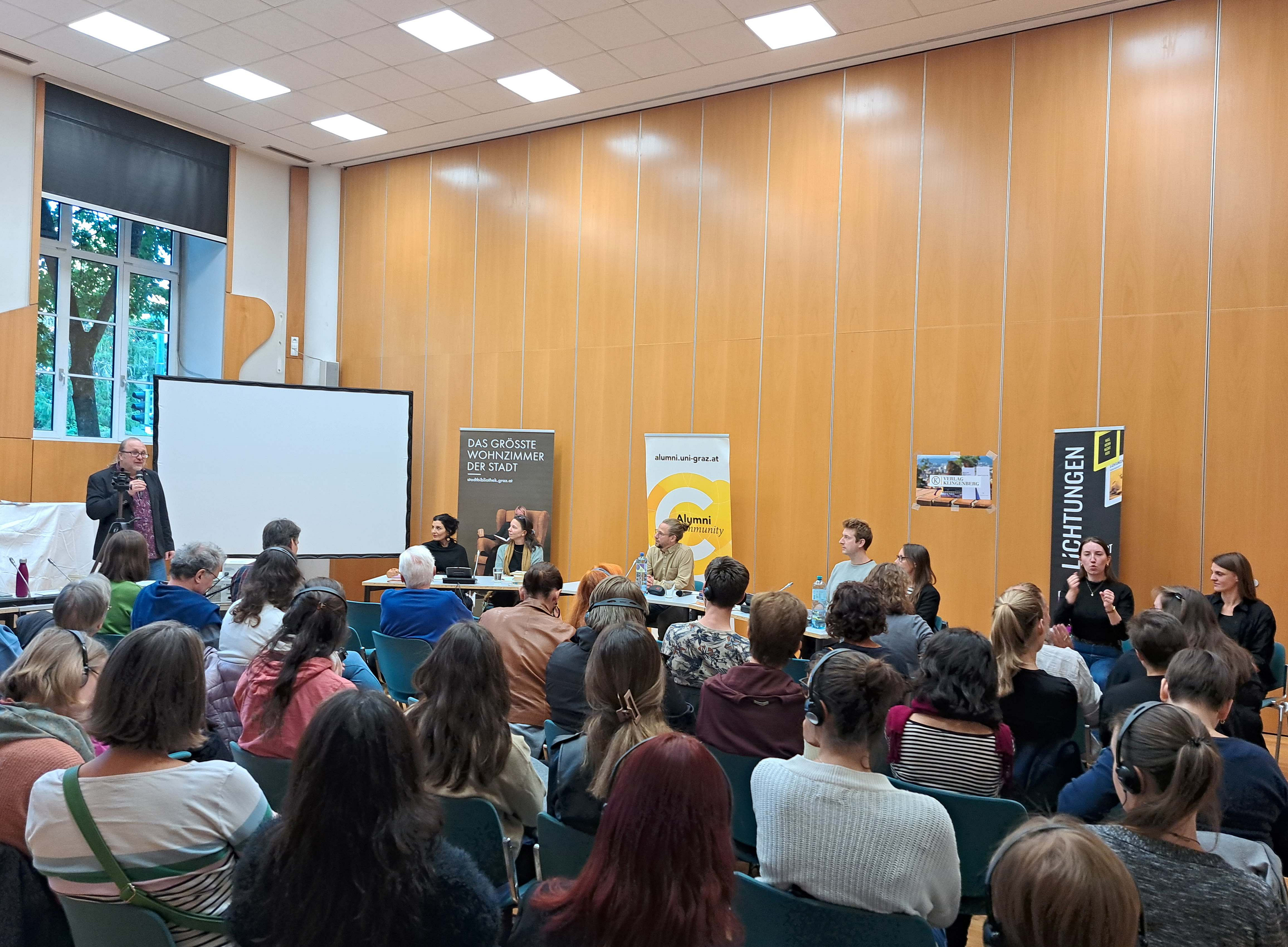 Personen sprechen vor vollbesetztem Saal; Blick von hinten 