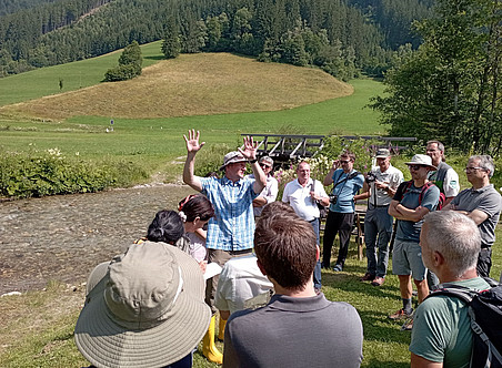 Eine Gruppe von Menschen in einer alpinen Umgebung, auf einem Außengelände in der Nähe von Wasser und grünen Hügeln mit Bäumen. Ein Mann steht mit dem Gesicht zur Kamera und winkt mit den Armen, während andere Männer in Polohemden oder T-Shirts und sommerlich gekleidete Frauen ihn anstarren. Er erklärt ihnen etwas über den Fluss und die Natur. 