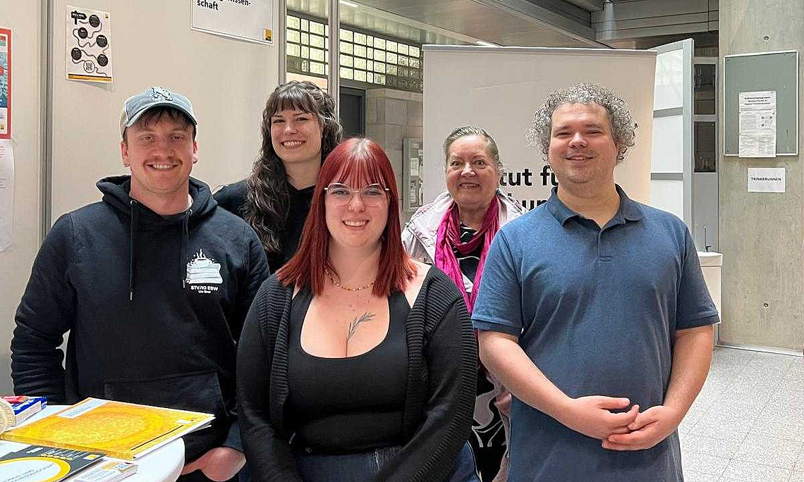 eine Professorin und 4 Studierende stehen vor einem Infostand mit Luftballons