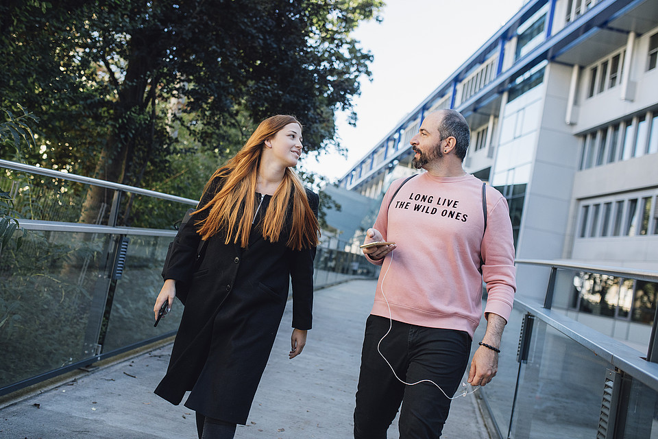 Zwei Studierende unterhalten sich, während sie auf einer schmalen Brücke am Campusgelände gehen