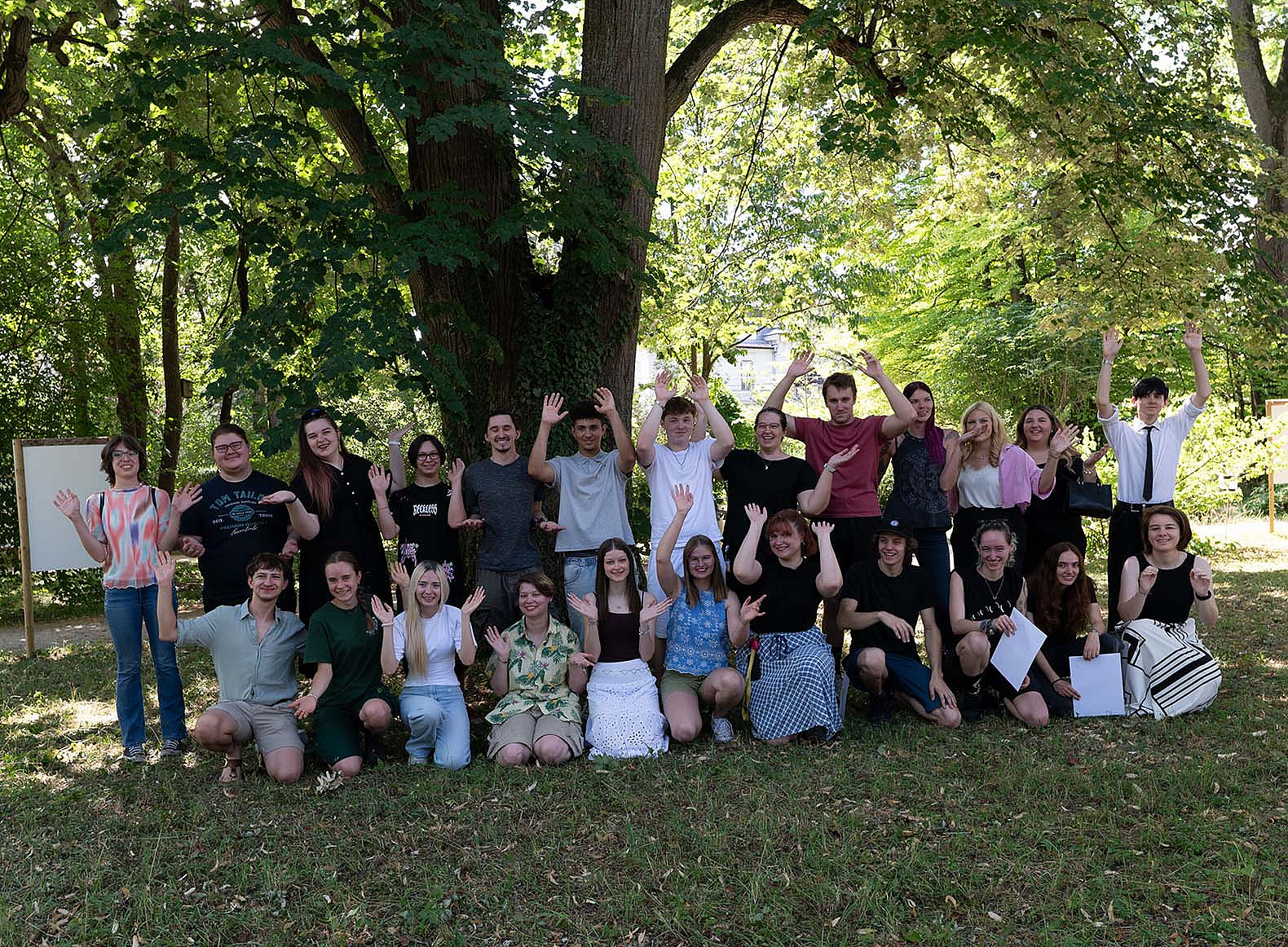 Gruppenfoto von 26 Lehrlingen bei der Abschlussfeier im Freien: Sie stehen und knien in zwei Reihen unter großen Bäumen und winken lachend in die Kamera. ©Uni Graz/Leljak