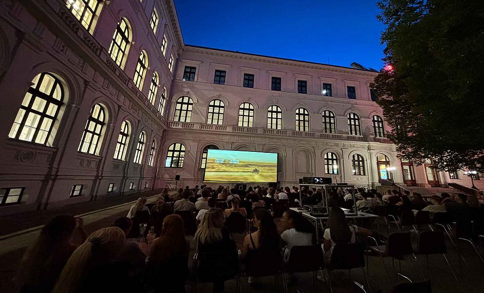 Viele junge Erwachsene sitzen abends auf Stühlen im Innenhof vor einer Leinwand und schauen einen Film; umgeben von einem historischen Gebäude.