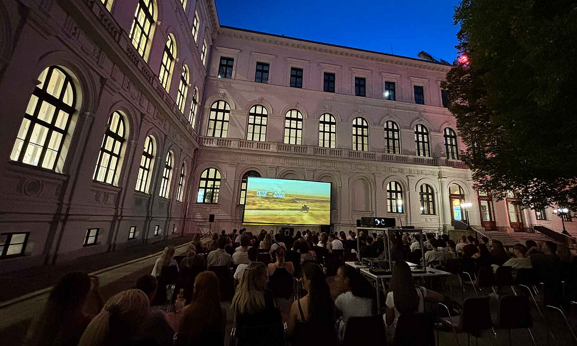 Viele junge Erwachsene sitzen abends auf Stühlen im Innenhof vor einer Leinwand und schauen einen Film; umgeben von einem historischen Gebäude.