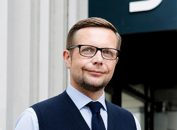 Mittelalter Mann mit Brille, kurz geschnittenem Haar, Hemd, Krawatte und dunkelblauer Weste steht vor einem modernen Gebäude mit großem Buchstaben. 
