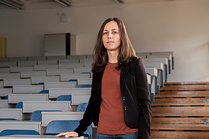 Photograph of a female professor standing in the middle of an empty university classroom, with blue and white seats. The shot is frontal, and the professor is wearing jeans, a black blazer, brown shoes, and an orange top.