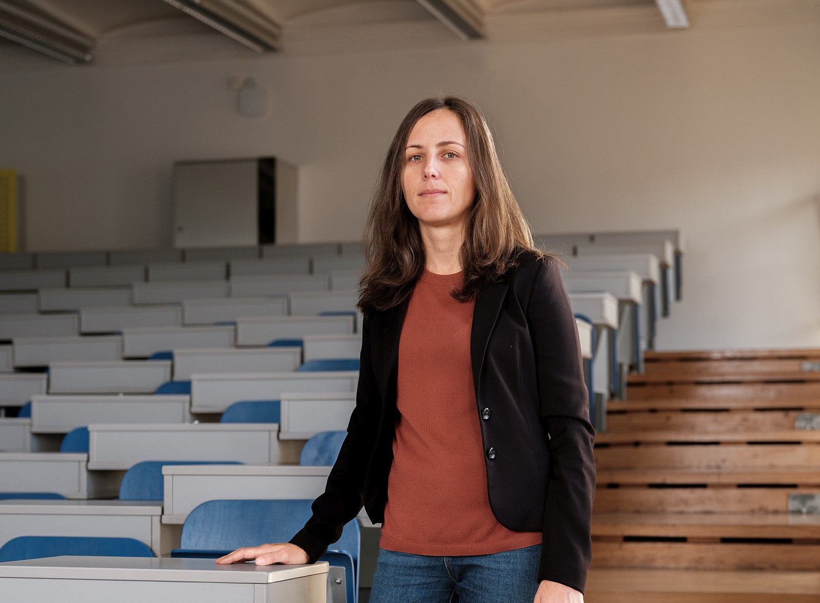 Photograph of a female professor standing in the middle of an empty university classroom, with blue and white seats. The shot is frontal, and the professor is wearing jeans, a black blazer, brown shoes, and an orange top. ©Manuel Schaffernak