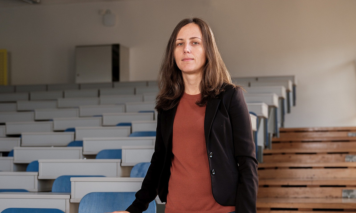 Foto einer Professorin, die in der Mitte eines leeren Universitätshörsaals mit blauen und weißen Stühlen steht. Die Aufnahme ist frontal, und die Professorin trägt Jeans, einen schwarzen Blazer, braune Schuhe und ein orangefarbenes Oberteil.